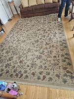Photo showing full view of large rectangular area rug with beige background, floral and grapevine patterns, laid on wooden floor in front of brown couch with cushions.