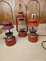 Three vintage red oil lanterns grouped together on a white surface against a wood-paneled wall. The middle lantern has a worn glass globe needing replacement.