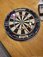Top-down view of the Winmau dartboard mounted on a wooden surface with six darts attached.