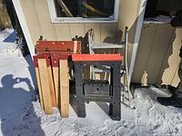 View of two wooden sawhorses with red tops, two black plastic sawhorses also with red tops, and an aluminum ladder in snow outside a building.