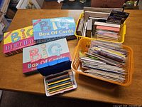 Wide shot showing two 'Big Box of Cards' boxed card sets for birthdays, two orange and yellow baskets filled with assorted greeting cards, and a tin of colored pencils on a wooden table.
