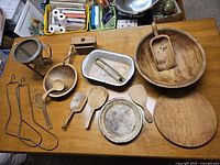Top-down view showing a collection of antique kitchen utensils and cookware including wooden bowls, scoops, cutting boards, a white shallow pan, and metal kitchen tools on a wooden table.