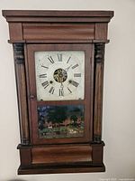 Front view of the antique grandmother wall clock showing the wooden case, clock face with Roman numerals, and painted glass panel beneath.