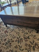 Closed view of solid walnut hope chest placed on patterned floral carpet, showing smooth rounded edges and visible wood grain.