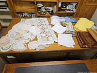 Overall view of white crocheted lace doilies, white linens, and folded brown, orange, and striped linen fabrics on wooden table.