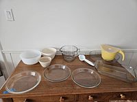 Full view of all eleven bakeware pieces displayed on wooden dresser, showing glass pie dishes, mixing bowls, batter dispenser, measuring spoon.