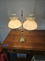 Photo of the front of the 1940's two armed brass desk lamp on wooden table, showing opaline glass shades illuminated.