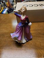 Figurine of woman in purple and pink dress holding fan to forehead, light blond hair curly, standing on table.