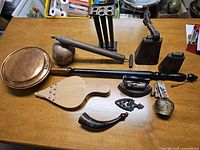 Photo of the full collection of 10 antique Canadiana items on a wooden table. Includes copper kettle, wooden and metal bellows, metal candle holder, irons, cowbells with straps.