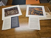 Two lithographs spread on a wooden table alongside accompanying descriptive text sheets. One depicts a birch forest scene and the other rolling hills.