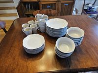 Full view of dinnerware set items on wooden table showing stacks of plates, bowls, and mugs arranged.