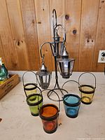 Photo showing full patio candle holder stand with three hanging lanterns and five colored glass candle cups arranged around it on a table.