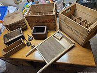 Wide view of eleven historic Canadiana wooden crates and tools on wooden table, showing beverage crates, cheese box, washboard, metal oil cans, and baskets.