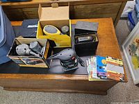 Wide shot of all sanding tools and accessories arranged on a wooden surface. Shows three boxed sanders and loose sanding papers.