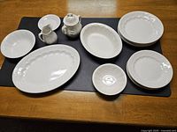 White ironstone dinnerware set on wooden table with black mat underneath displaying plates, bowls, serving platters, creamer, and sugar bowl.