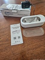 White Kunphy ultrasonic cleaner with clear lid, user manual, power adapter, and box on wooden table.
