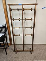 Full front view of the tall wooden bobbin/spool rack standing on floor against a wall showing its structure of vertical wooden supports and horizontal metal rods with wooden bobbins mounted.