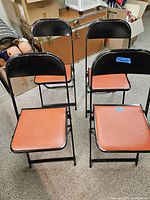 Four vintage metal folding chairs arranged in two rows showing black metal frames and orange seats in a carpeted room with various background items.
