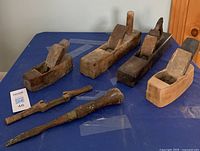 Front view of the six antique woodworking tools, showing five wooden hand planes and one wooden-handled screwdriver on a blue surface.