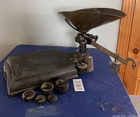 Full view of the cast iron mechanical weight scale with weighing pan, assorted cast iron weights displayed in front, and storage box to the side.