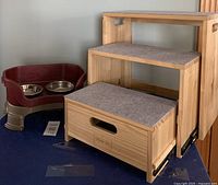 View of the dog stairs and dog feeder showing the wood stairs with felt tops next to the maroon and tan feeder with metal bowls.