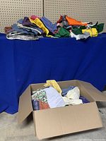 Photo showing multiple piles of assorted fabric pieces in various colors and patterns stacked on a blue draped surface with additional fabrics inside a cardboard box.