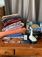 Stack of various folded fabrics in multiple colors and patterns with handheld steamer and some sewing notions placed on wooden dresser
