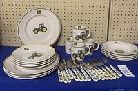 Full set of John Deere dinnerware and cutlery displayed, showing plates, bowls, mugs and matching utensils on blue cloth with beige pegboard background.