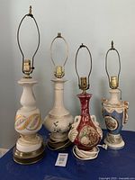 Four decorative ceramic and metal table lamps on a blue surface. No lamp shades included. The lamps vary in color and decoration, with visible cords and lamp holders.