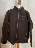 Front view of the black Milwaukee heated jacket with zipper closed, showing Milwaukee logo on chest and a small zippered pocket with heating indicator.