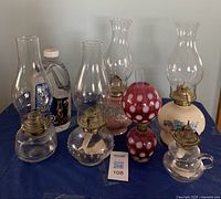 Photo showing six assorted oil lamps with clear glass chimneys and a new plastic jug of lamp oil on a blue cloth background.