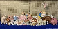 Wide view showing full collection of pig-themed items on blue cloth, including mugs, figurines, dolls, piggy banks, and a large brown ceramic pig on the right.