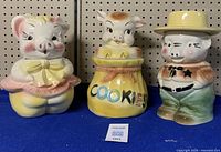 Three pig cookie jars side by side on blue shelf against pegboard backdrop