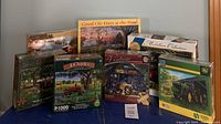 Seven sealed or nearly sealed jigsaw puzzles arranged on a flat surface showing various scenic and vehicle themes including farm tractors, vintage cars, and rural landscapes.