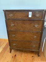 Front view of tall wooden dresser showing four drawers, metal oval drawer pulls, and decorative vertical reeded columns on corners. Wood surface shows wear and finish imperfections.