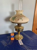 Full view of the vintage Aladdin brass oil lamp with daisy patterned glass shade and clear chimney, showing included extra mantle box beside it on table.