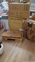 Photo showing several stacked boxes including a bread maker, food warming mat, warming tray, and woodgrain-patterned box on floor.