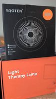 Boxes showing YAOTEN brand, orange box labeled Light Therapy Lamp with image of the rectangular lamp panel and adjustable stand, black box labeled LED Multi-Function with image of circular fan blade and specifications.