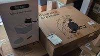 Photo showing both the boxed Rizzari Smart Pet Feeder and the Outdoor Cat House with a cartoon cat illustration on each box.