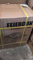 Box containing a Feierdun stepper machine secured with yellow straps, showing brand and packaging illustration.