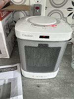 White Lasko ceramic heater plugged into wall outlet with control dial set to 'low' and red indicator light on. Original box and instruction manual visible beside heater.