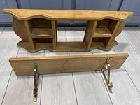 Two wooden wall shelves shown from front and side views on a tiled floor. One is a multi-compartment wood shelf with curved top edges; the other is a plank shelf with brass hanging brackets.