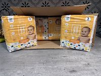 Four sealed packs of Hello Bello diapers size 3 placed partially inside and outside a cardboard box on a gray surface.
