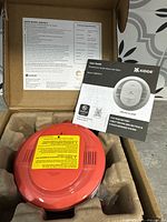Kidde smoke alarm front view showing the red circular housing with ventilation slots and attached warning label, along with user guide documentation visible inside the packaging box.