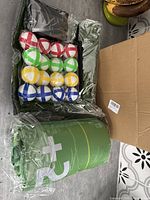 Top view of multiple colored golf balls and green cylindrical target, both in plastic packaging, alongside black carry case