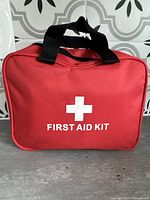 Closed red First Aid Kit bag with black handles and white cross logo text on front