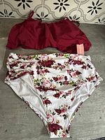 Photo of both pieces of the two-piece bathing suit lying flat, showing the dark red ruffled top and the floral high-waisted bottom with tags attached.