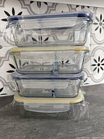 Stack of 4 clear glass casserole dishes, two with internal dividers, two without, with blue and yellow silicone-sealed airtight lids, all wrapped in plastic.