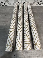 Four new rolls of peel and stick wallpaper with gold and white geometric pattern on a grey background surface.