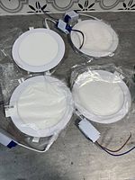 Four white round LED pot lights with drivers, some wrapped in plastic, shown on a concrete floor.